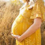 donna incinta con vestito lungo giallo tra spighe di grano si tocca la pancia